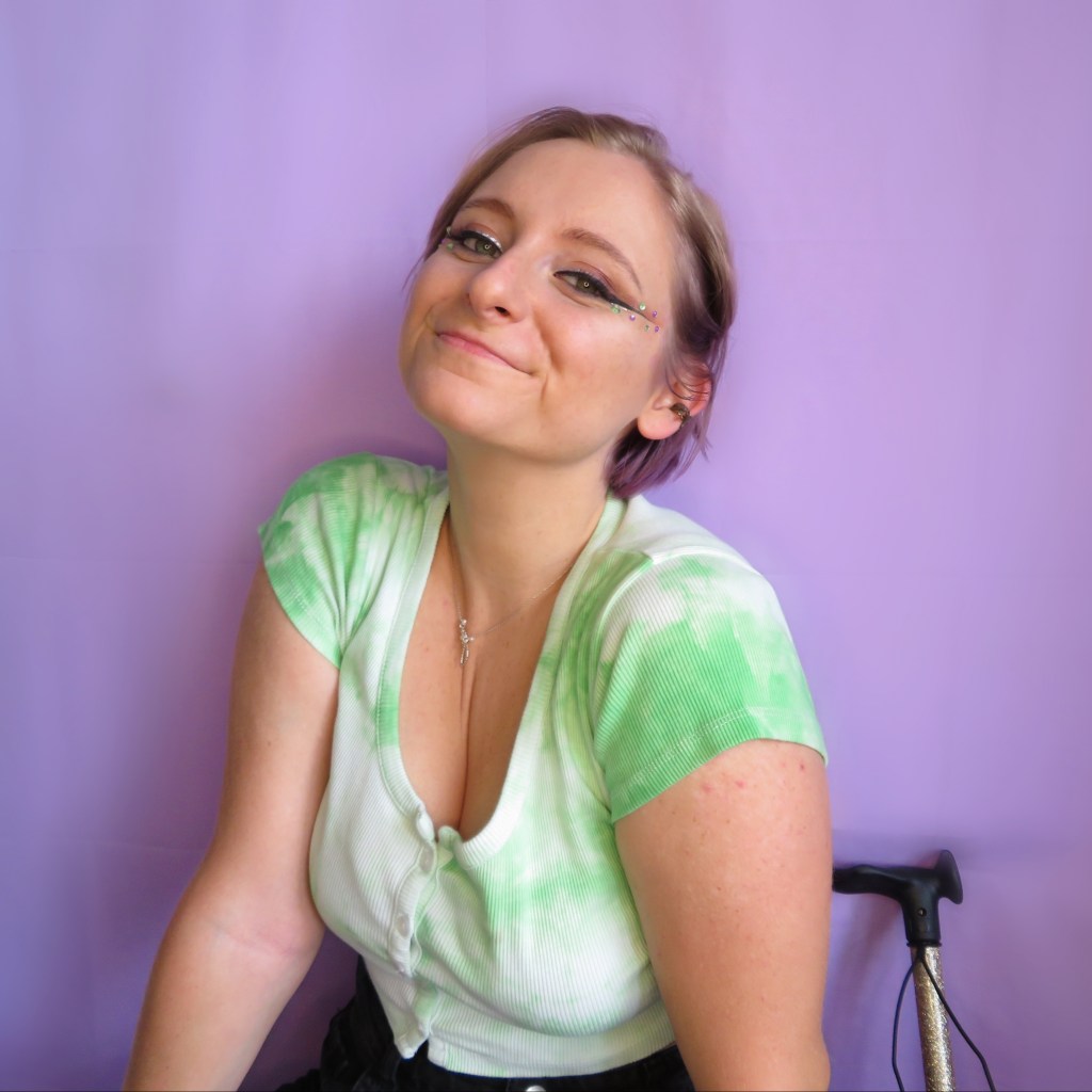 Charlotte, a white woman in her 20s with short blonde hair, smiling at the camera. They are wearing a green top and there's a gold walking stick behind her.